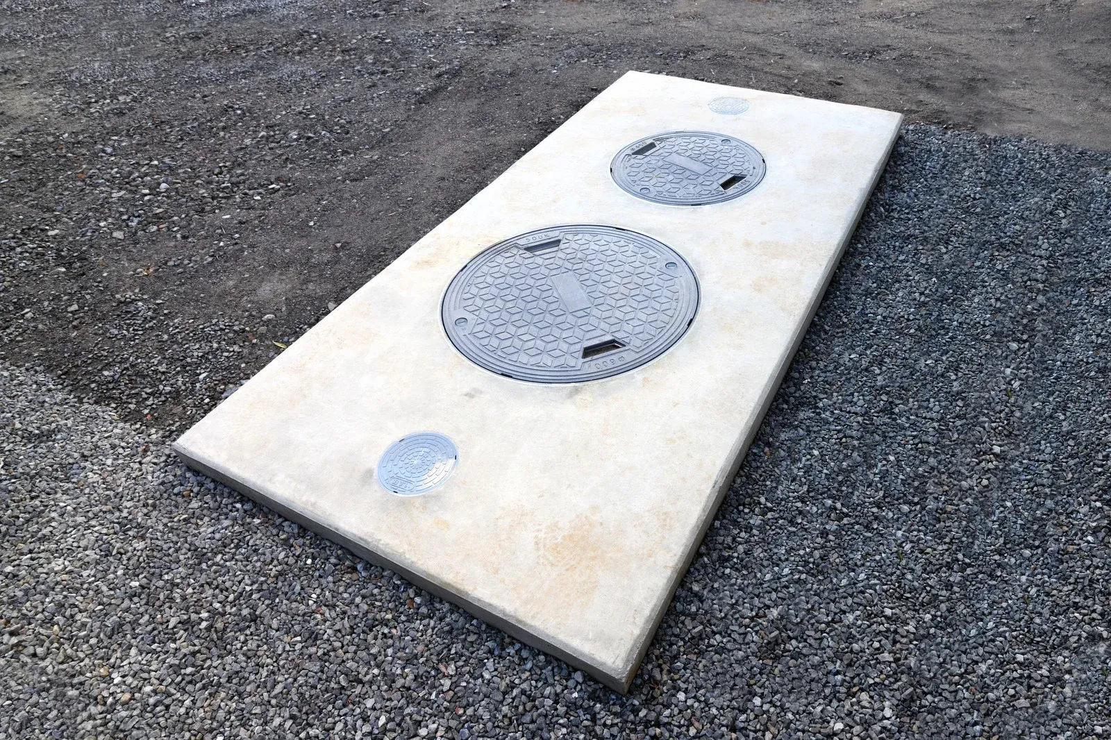 Concrete slab with two circular access covers and a smaller circular access point, set on gravel.