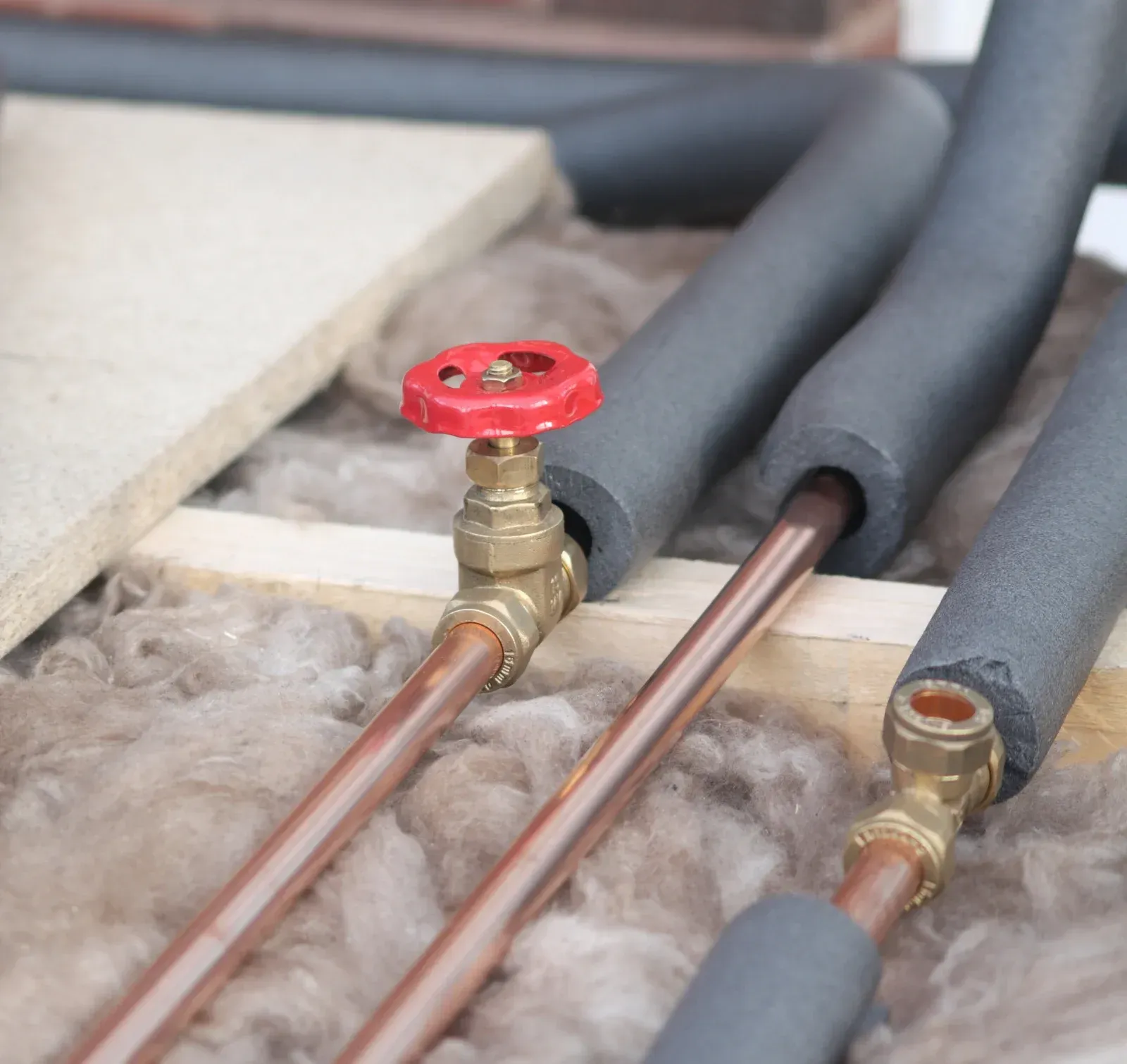 Copper pipes with insulation and a red valve, set in wooden frame with loose insulation.