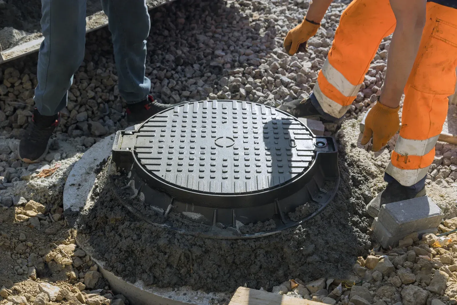 Workers installing a dark, round manhole cover in concrete, wearing work clothes and gloves.
