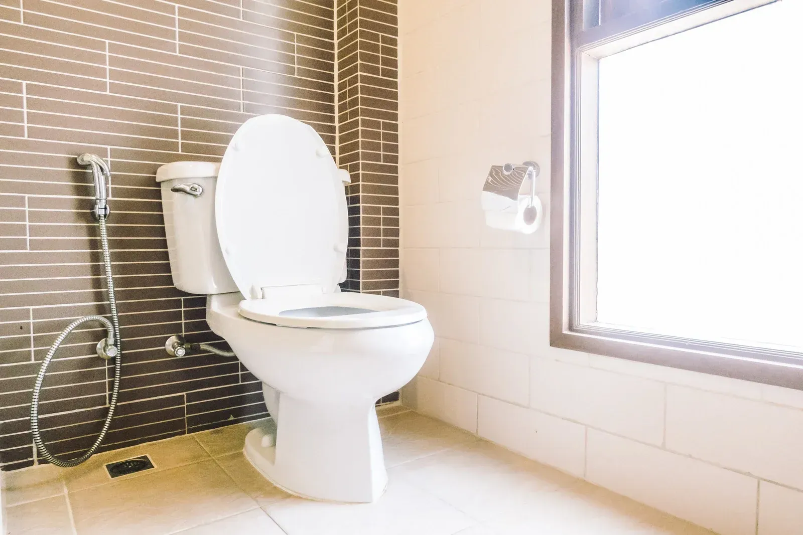 White toilet in a bathroom with brown and white tiles. A window is in the background.