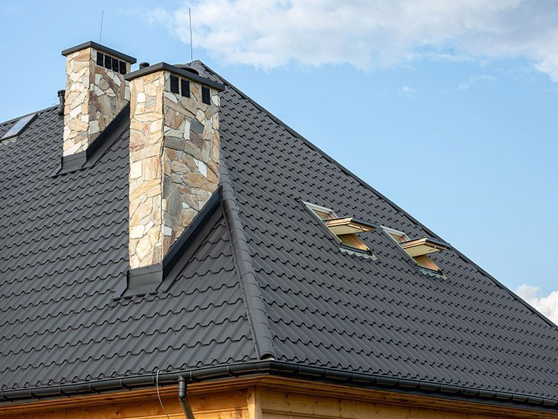 A dark gray metal roof with two stone chimneys and two open skylights against a blue sky.