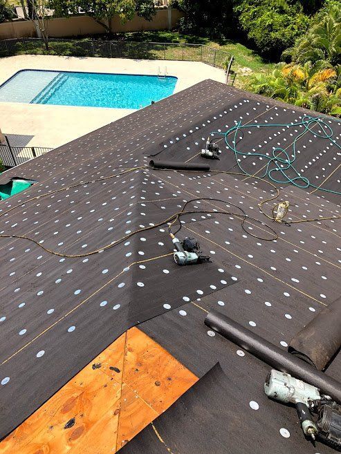 A roof under construction covered in black underlayment with fastening marks, next to a swimming pool in a sunny yard.