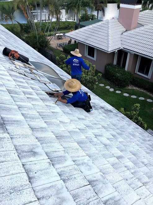 Two workers in blue shirts and straw hats repair a white-tiled roof on a sunny day.