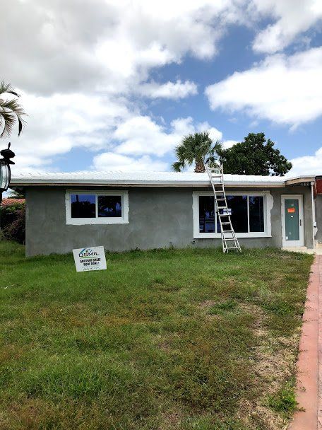 A gray single-story house under a blue, cloudy sky, with new white window frames and a ladder leaning against the roof.