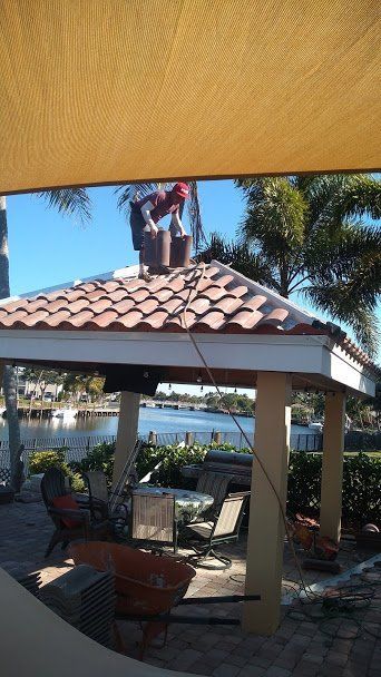 A person working on the tiled roof of an outdoor gazebo near a waterfront, with a patio set visible underneath.
