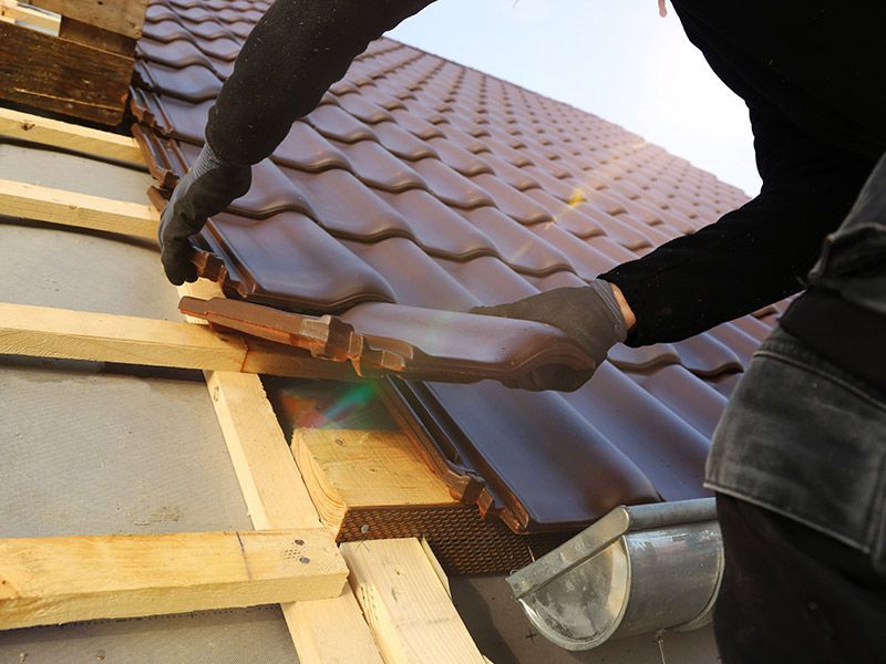 A person in gloves installs dark brown roof tiles onto a wooden frame on a rooftop.