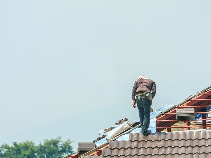 A worker in a brown shirt and dark pants stands on a partially tiled roof, working on a wooden frame against a blue sky.