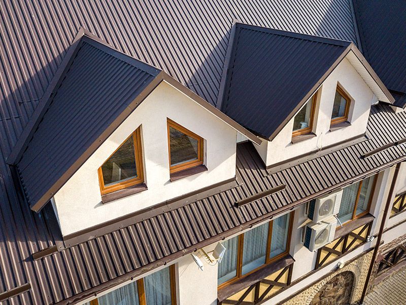 High-angle view of a building roof with two gabled dormer windows, featuring brown metal roofing and off-white walls.