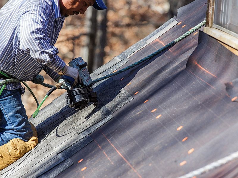 A worker in a striped shirt uses a pneumatic nail gun to install shingles on a sloped residential roof.