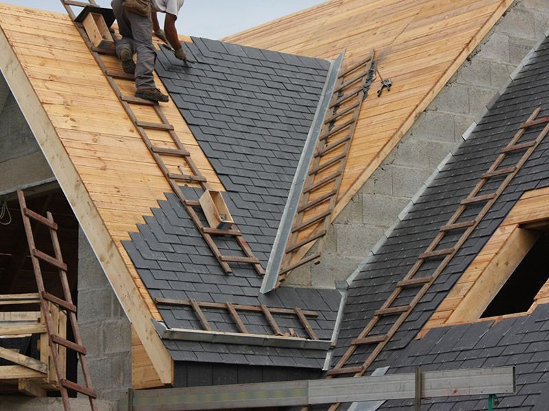 A construction worker installs dark shingles on a multi-pitched roof with wooden framing and metal flashing in a valley.
