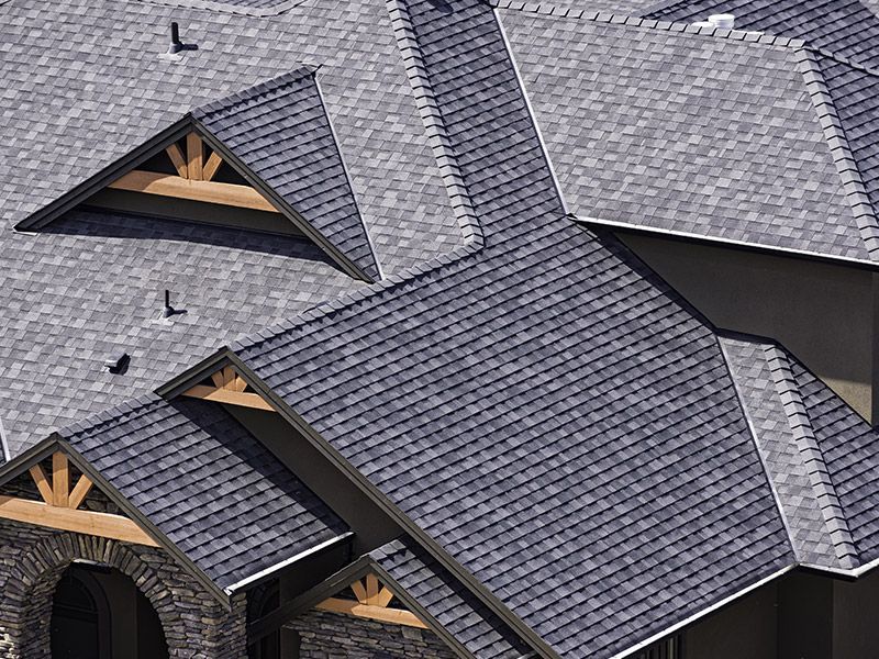 High-angle shot of a gray shingled roof featuring multiple gables with exposed timber beams and stone accents.