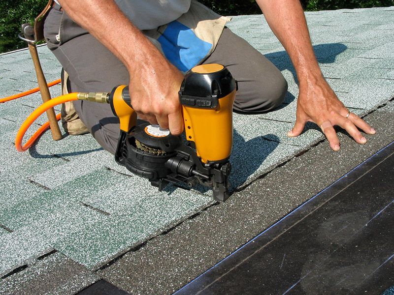 A person uses a yellow pneumatic nail gun to install grey asphalt shingles on a residential roof.