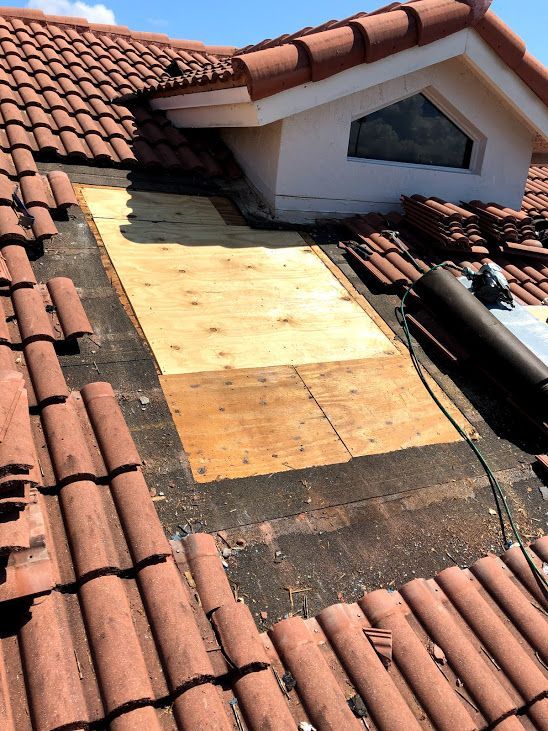A section of red tiled roof under repair, featuring new plywood decking installed against a white dormer window.