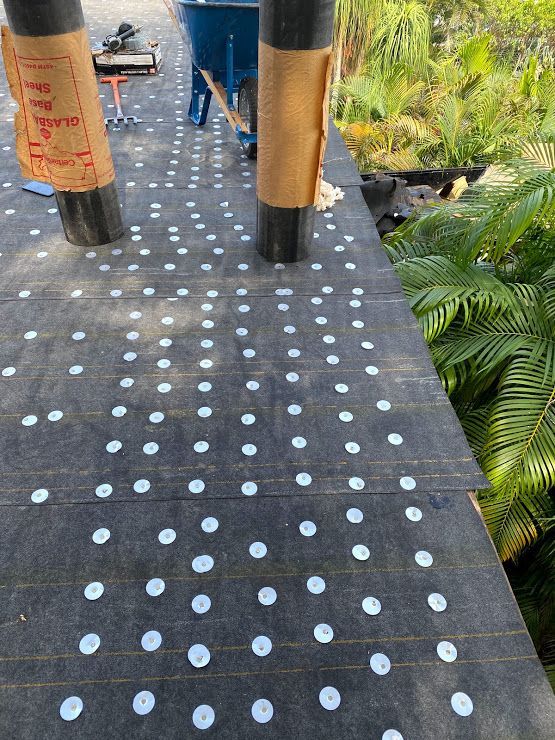 A black roof underlayment covered in a grid of white circular fasteners, with two wrapped support posts and a wheelbarrow.