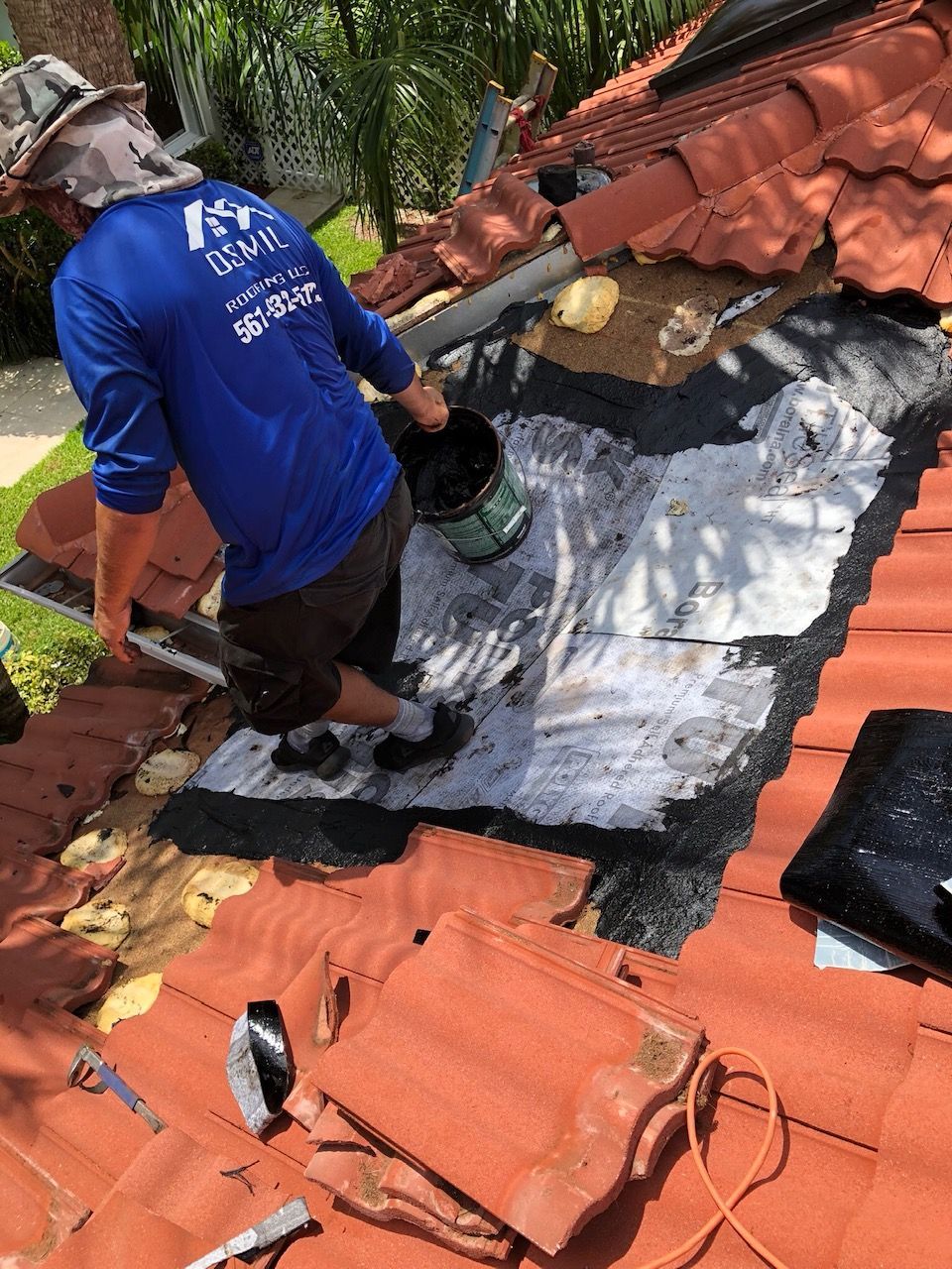 A worker in a blue shirt applies sealant to a tiled roof with missing tiles, exposing the underlayment underneath.