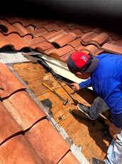 A worker in a blue shirt and red cap repairs a section of a terracotta tile roof, using a hammer on the wooden underlayment.