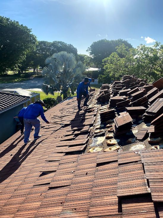 Two workers in blue uniforms remove clay roof tiles from a residential roof on a sunny day.