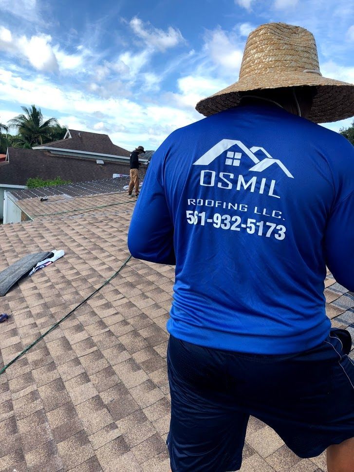 A worker wearing a blue branded long-sleeve shirt and straw hat stands on a residential roof during a repair.