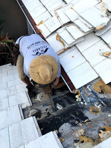 A worker in a straw hat and grey shirt repairs a white tile roof, removing tiles to access the underlayment.