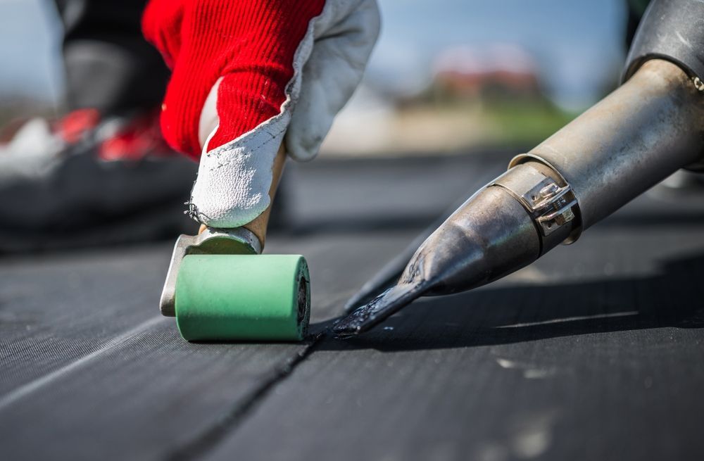 A person wearing red gloves uses a heat gun and a green roller to seal a seam on a black roofing membrane.