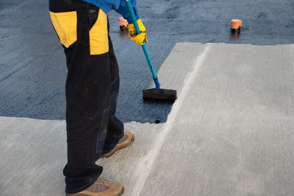 A worker in work pants and gloves uses a roller to apply black sealant to a gray concrete surface.