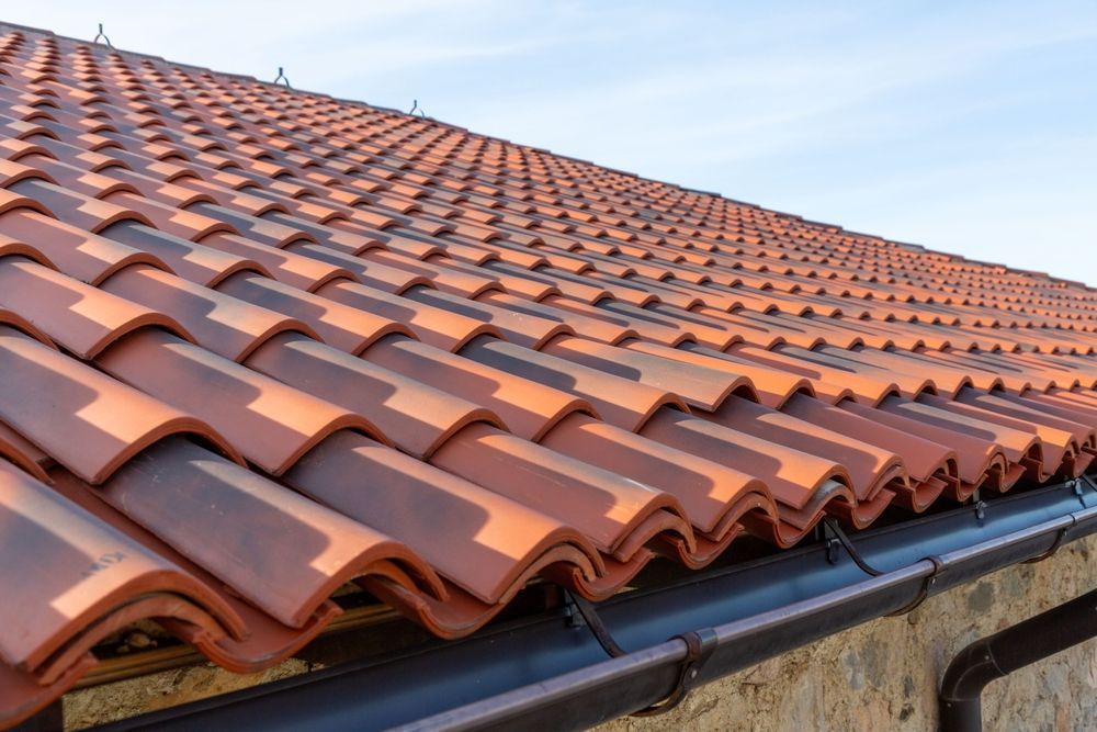 Close-up of a terracotta-colored, wavy roof tile pattern on a building with a dark gutter against a pale sky.
