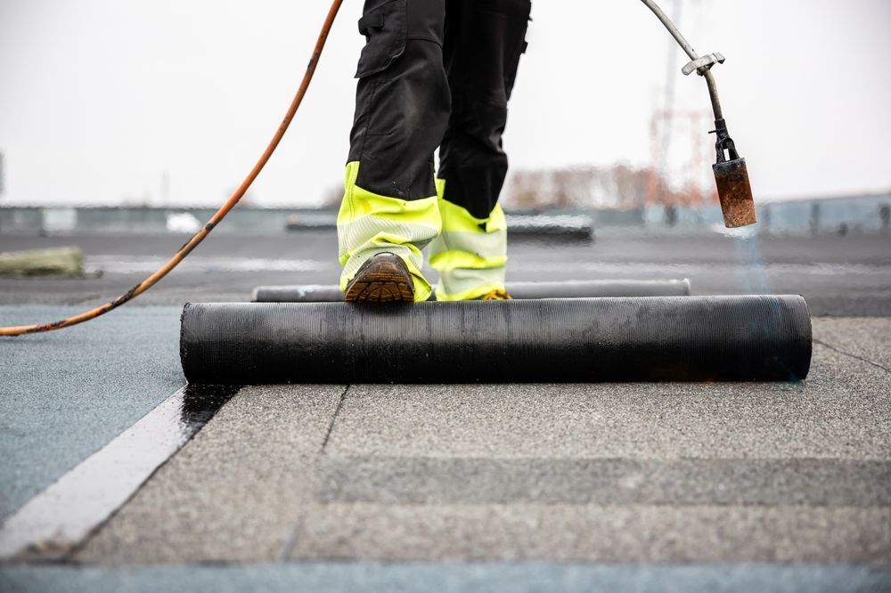 A worker in high-visibility yellow trousers uses a propane torch to install roofing material on a flat surface.