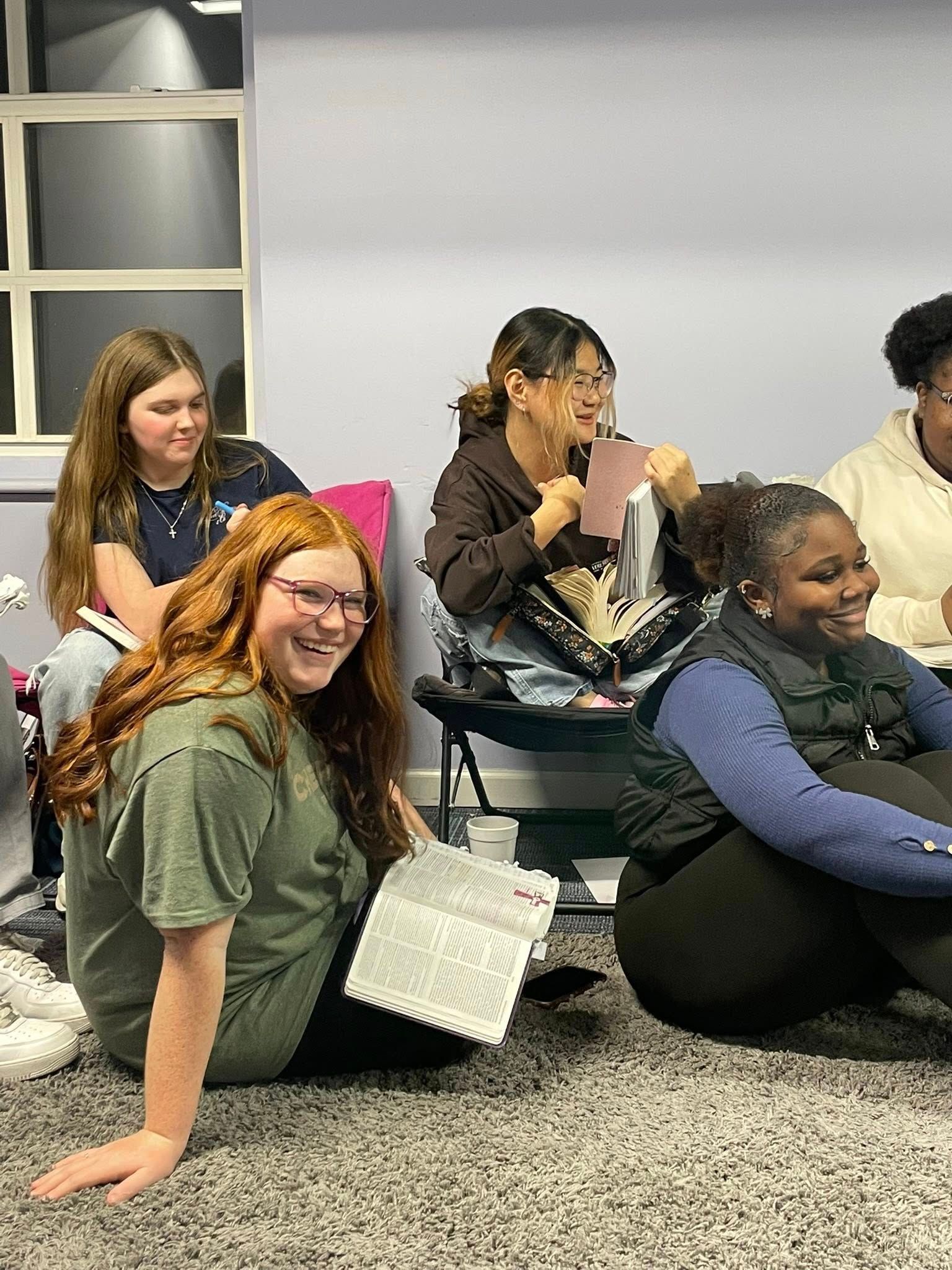 People sitting on the floor in a classroom, smiling and chatting with notebooks nearby