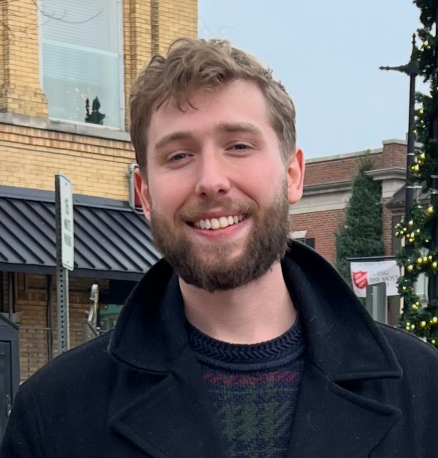 Smiling bearded person in a dark coat standing outdoors on a city street