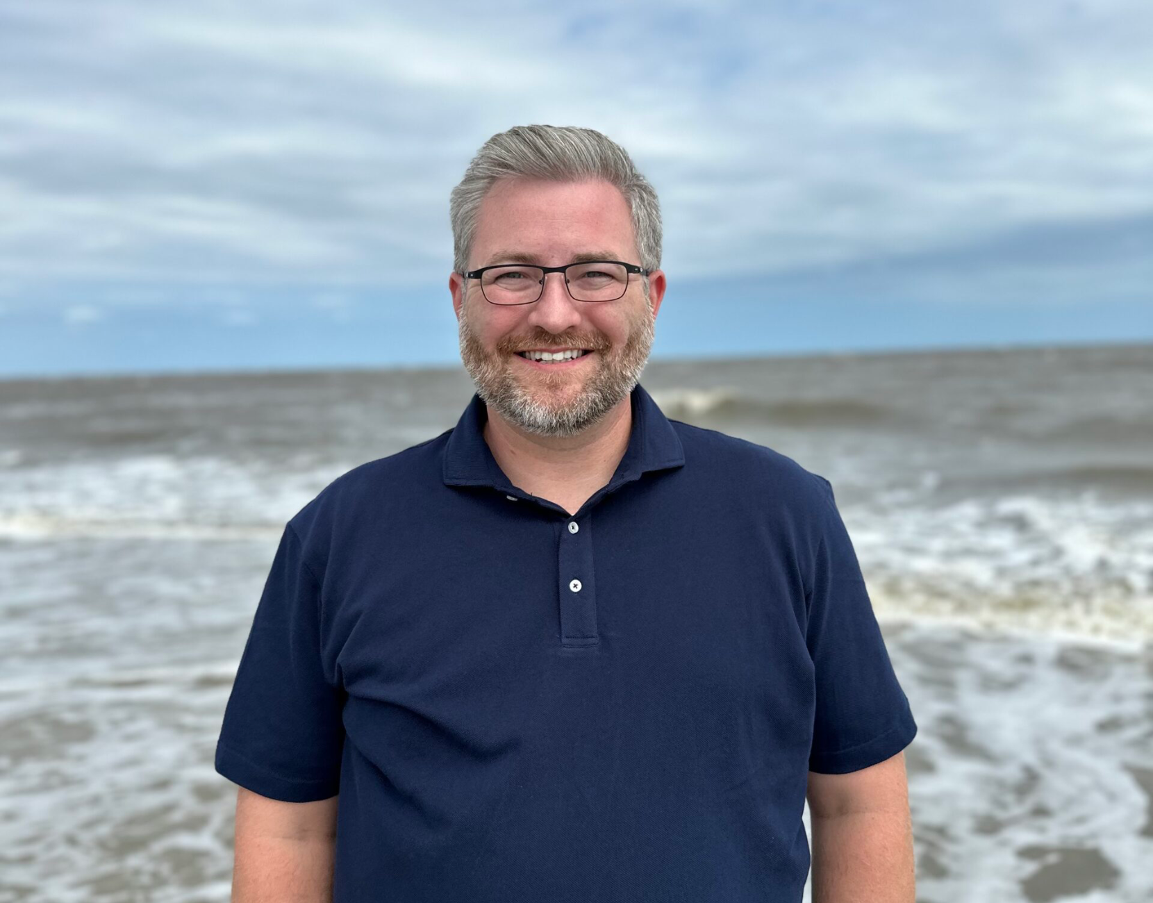 Smiling man in a navy polo standing at a windy beach with waves and cloudy sky