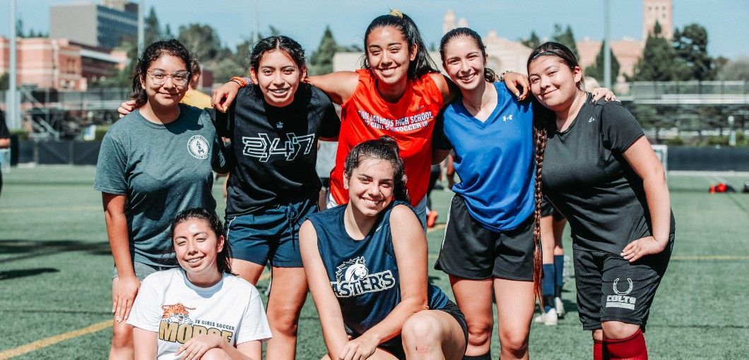 Seven teammates posing on a soccer field, smiling in jerseys and uniforms.