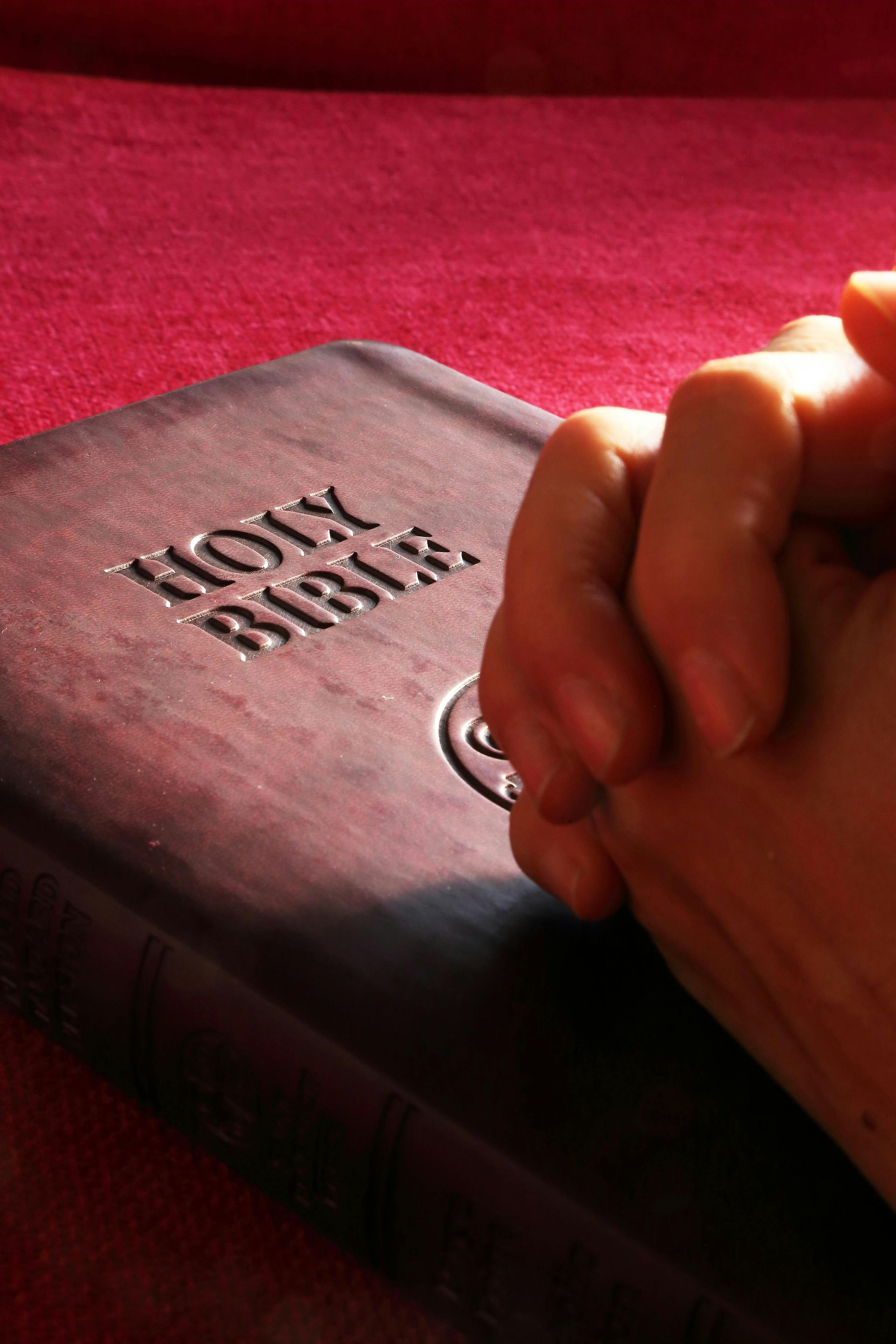 Hands clasped in prayer over a Bible on a red surface