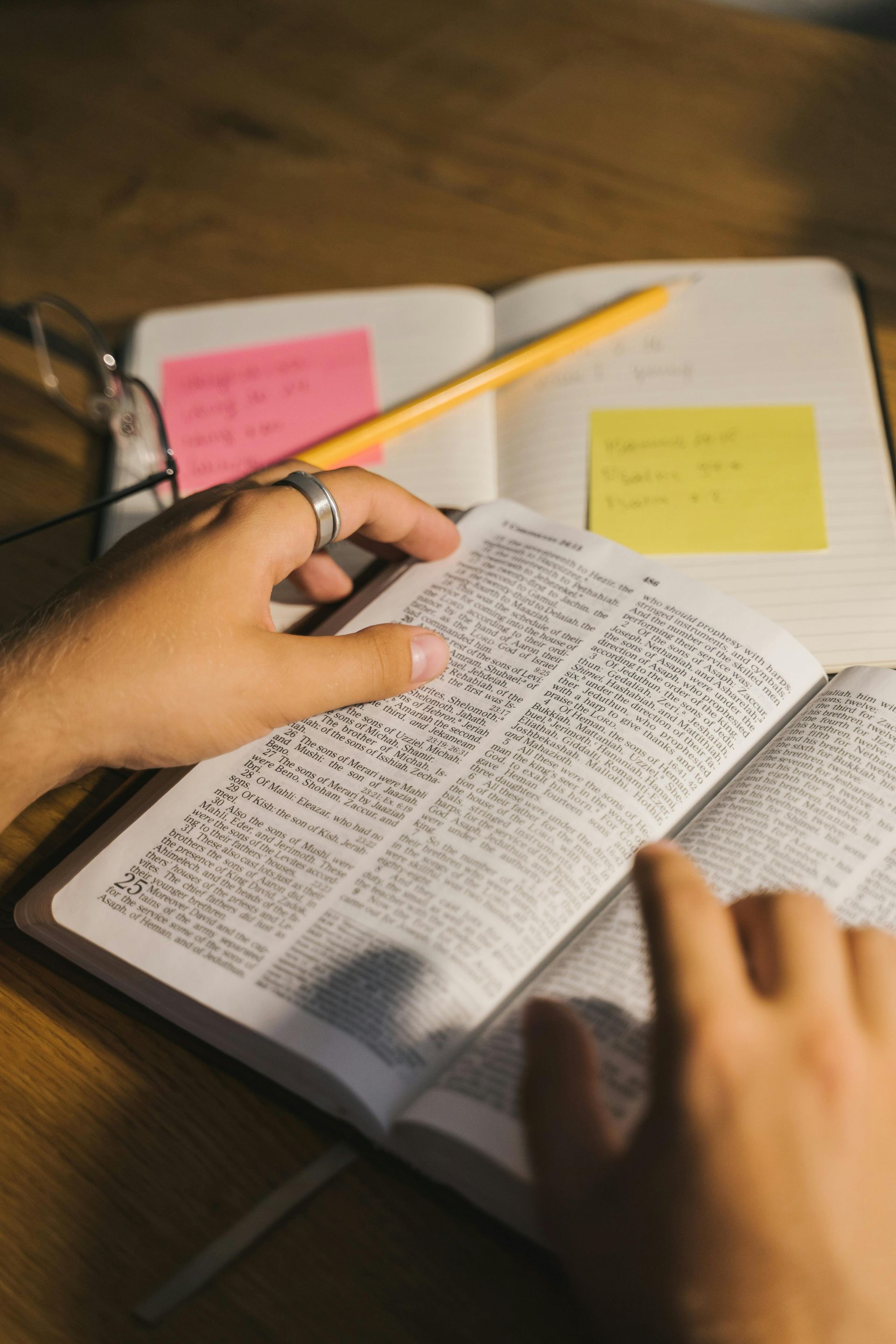 Hands taking notes on an open notebook and small Bible with sticky notes and pencil nearby