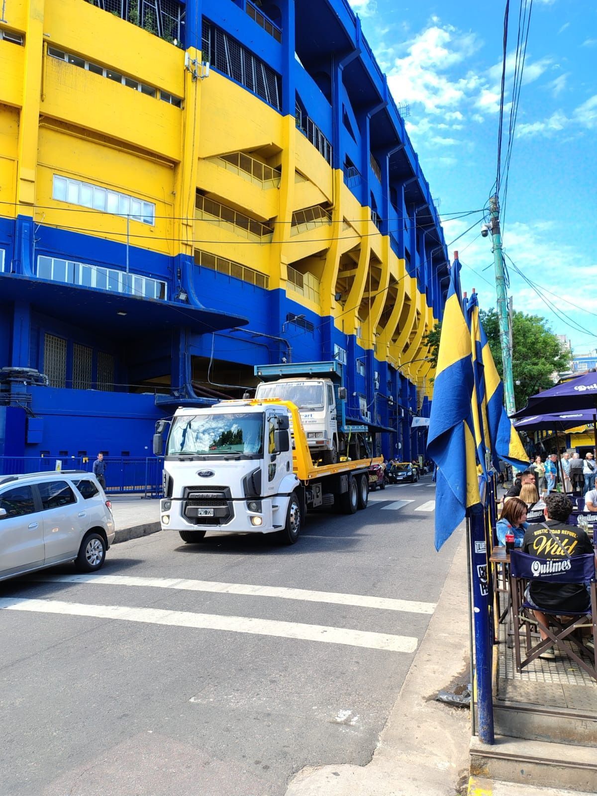 Camión circulando por la calle al lado de un estadio azul y amarillo con gente y banderas visibles.