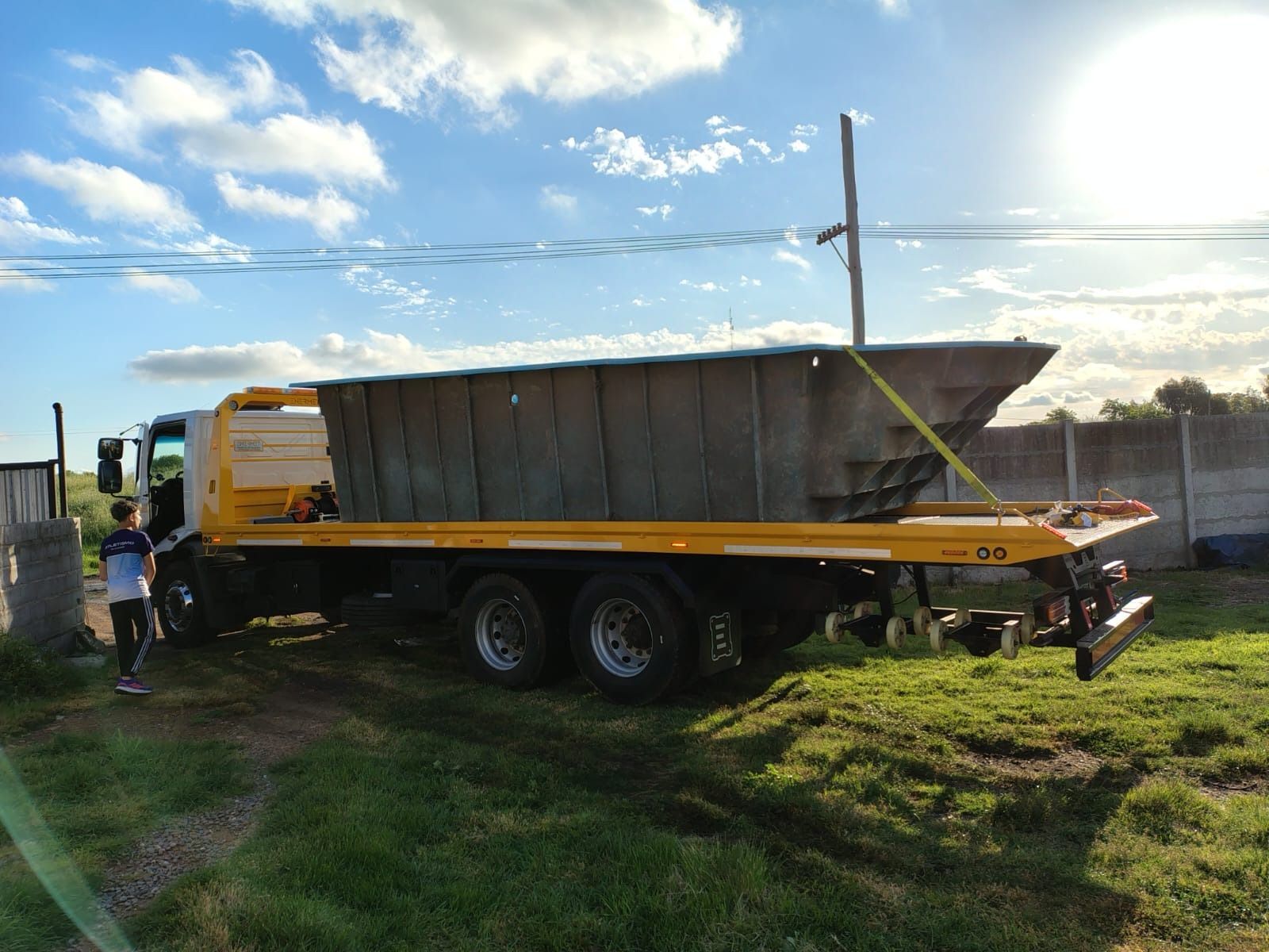 Un camión de plataforma transporta un objeto grande, similar a un contenedor. Una persona se encuentra cerca, en una zona verde bajo un cielo soleado.