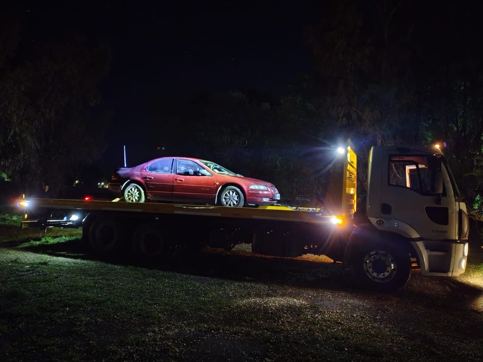 Un coche rojo es remolcado por una grúa de plataforma de noche. Luces brillantes iluminan la escena.