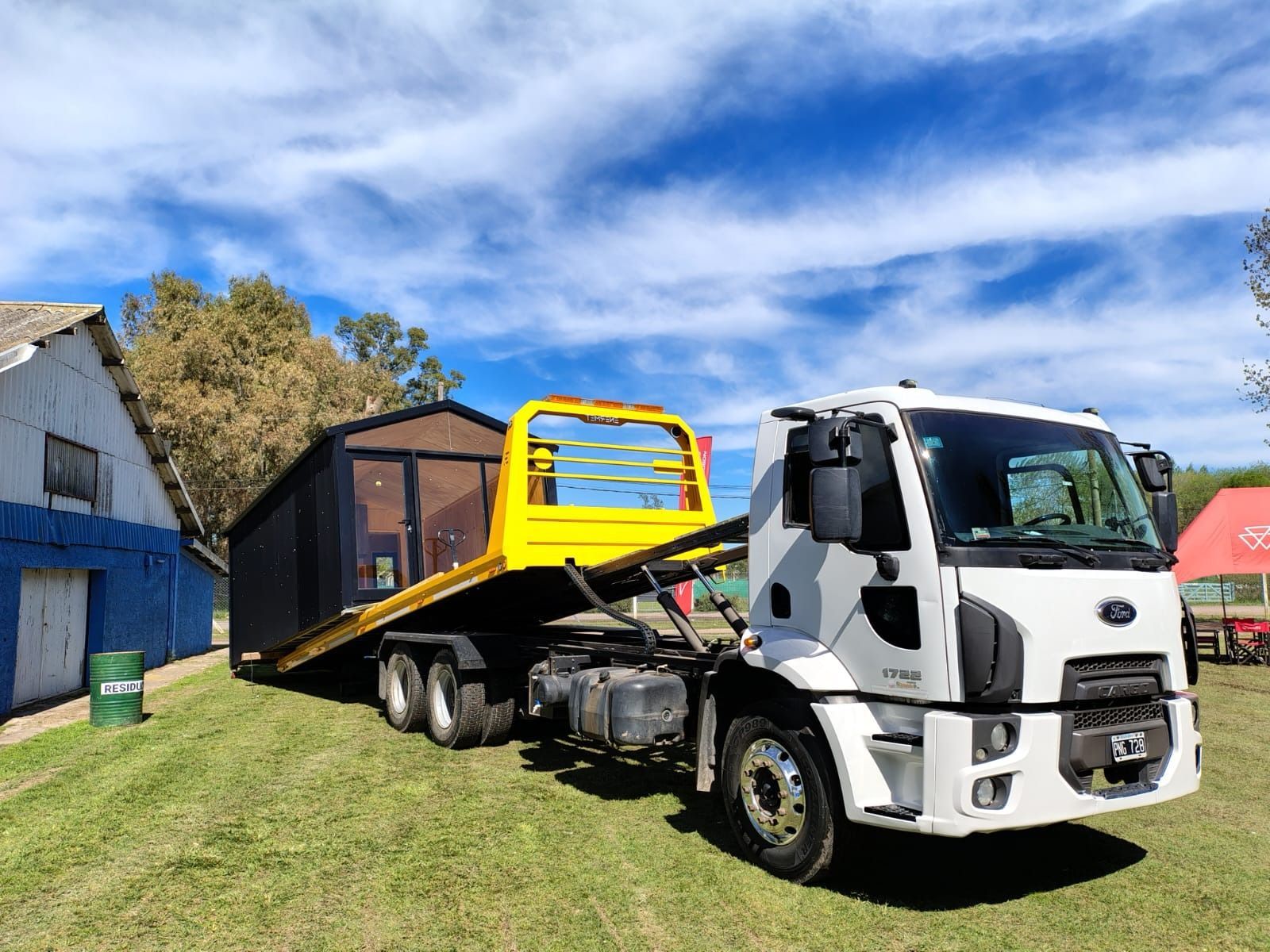 Un camión Ford blanco con plataforma amarilla transporta una caja negra sobre una zona cubierta de hierba, bajo un cielo azul.