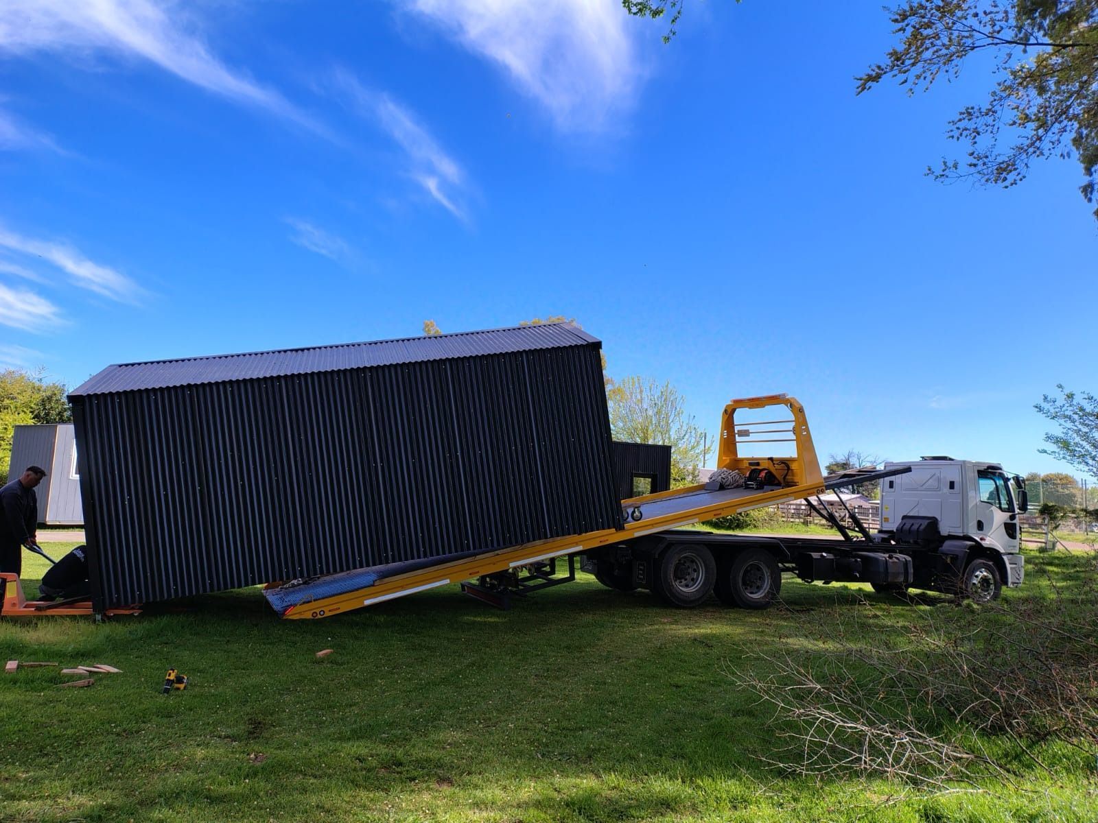 Un cobertizo oscuro siendo cargado en un camión de plataforma sobre césped verde bajo un cielo azul.