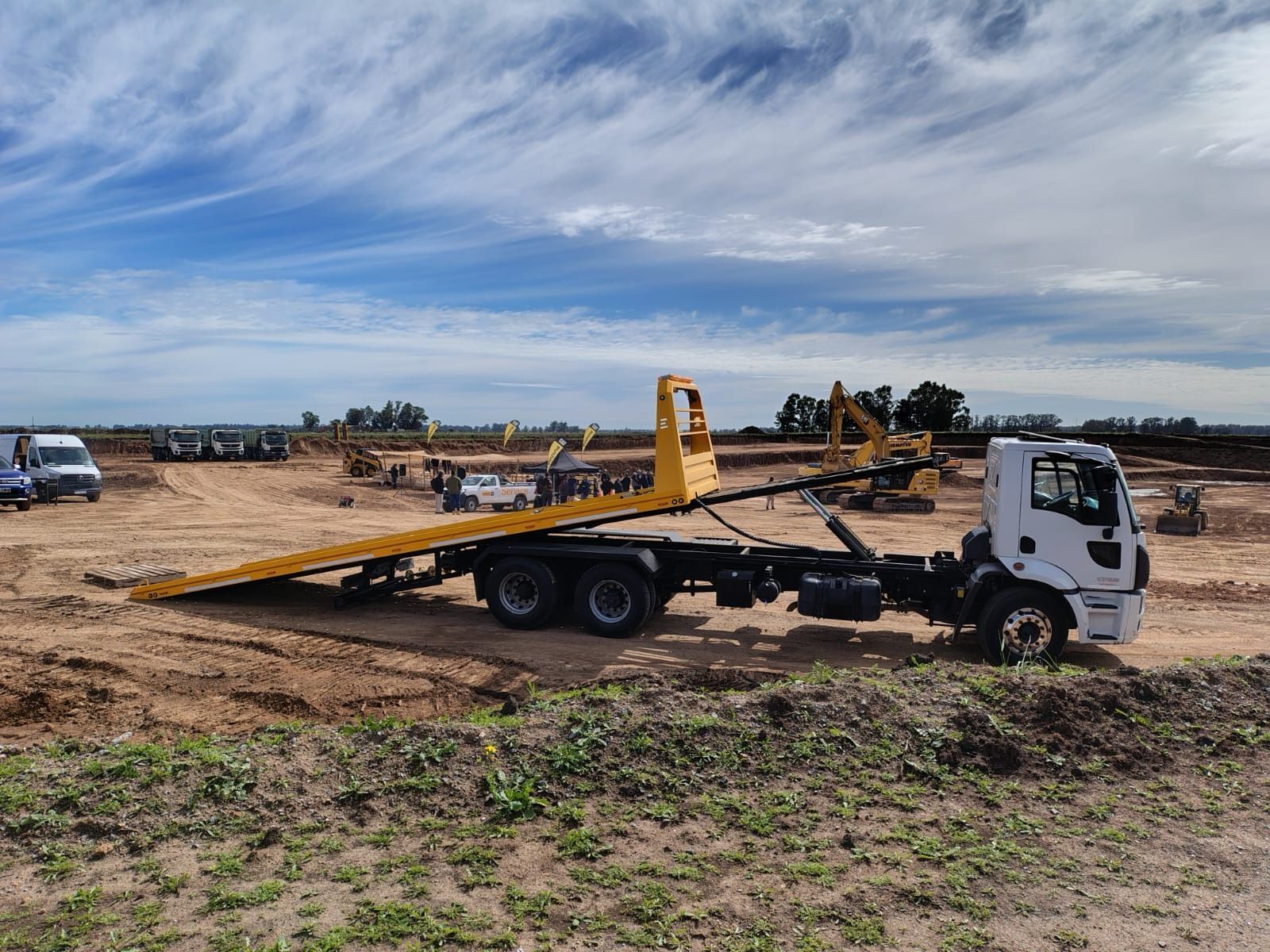 Camión de plataforma sobre tierra, caja amarilla, cabina blanca, al aire libre.
