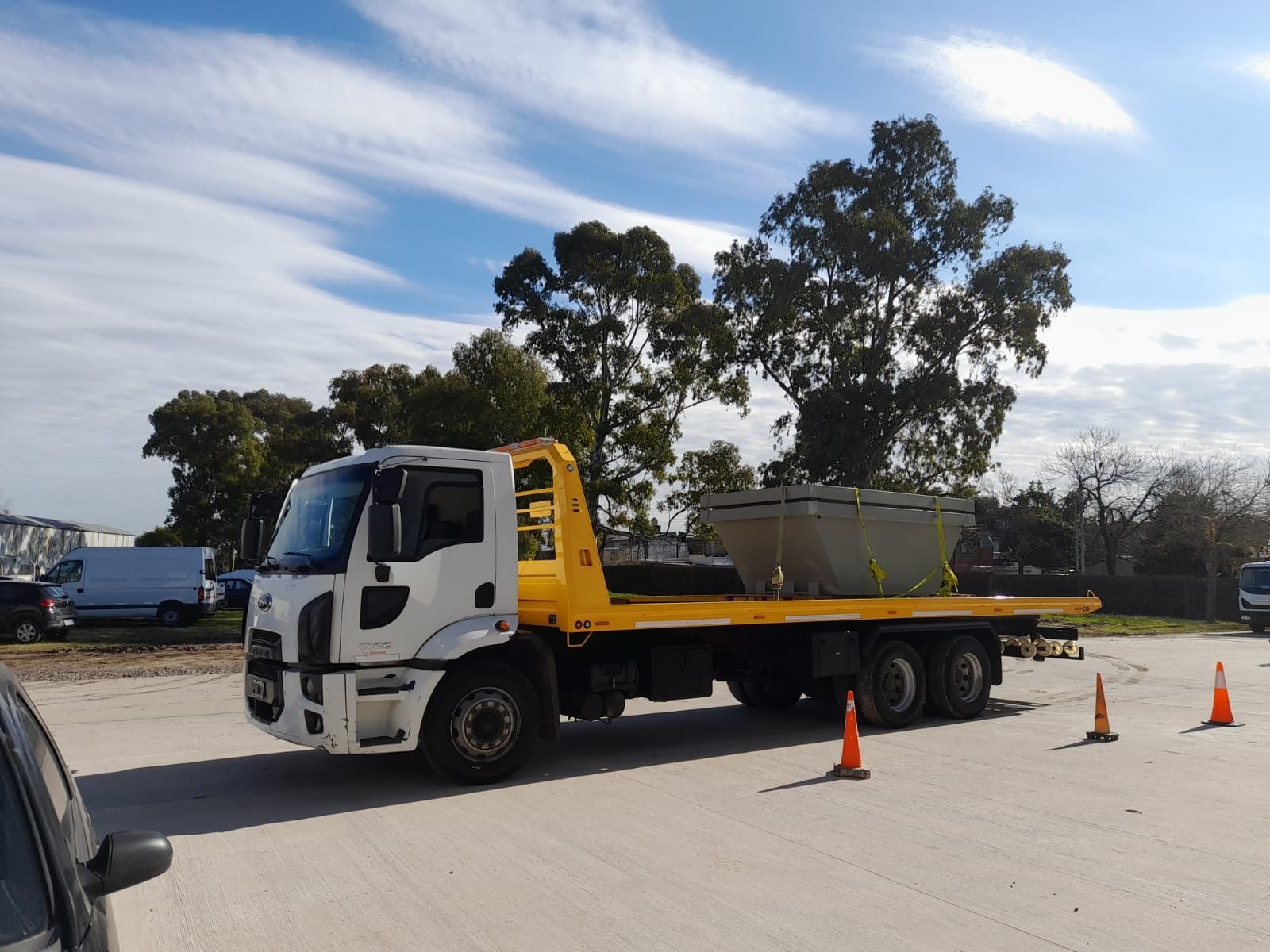 Grúa con plataforma que transporta un gran objeto de hormigón, al aire libre. Camión amarillo y blanco, cielo despejado.