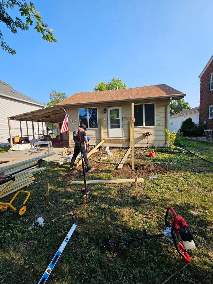 Man building a structure near a small house with a flag. Sunny day, grassy yard, tools scattered.