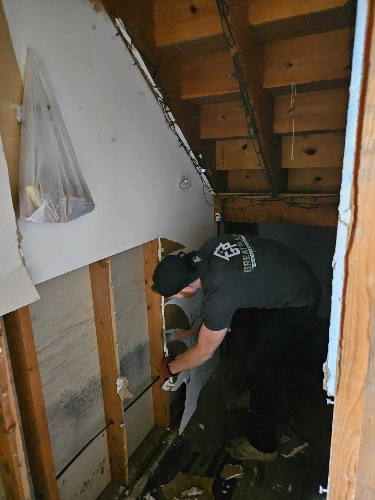 Man removing drywall from a wall in a construction area. Wooden beams and framing are visible.