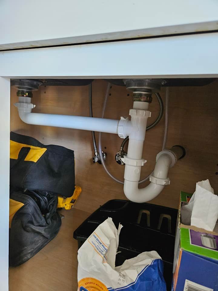 Under-sink plumbing with white pipes and a U-shaped trap connecting two sink drains, inside a white cabinet.
