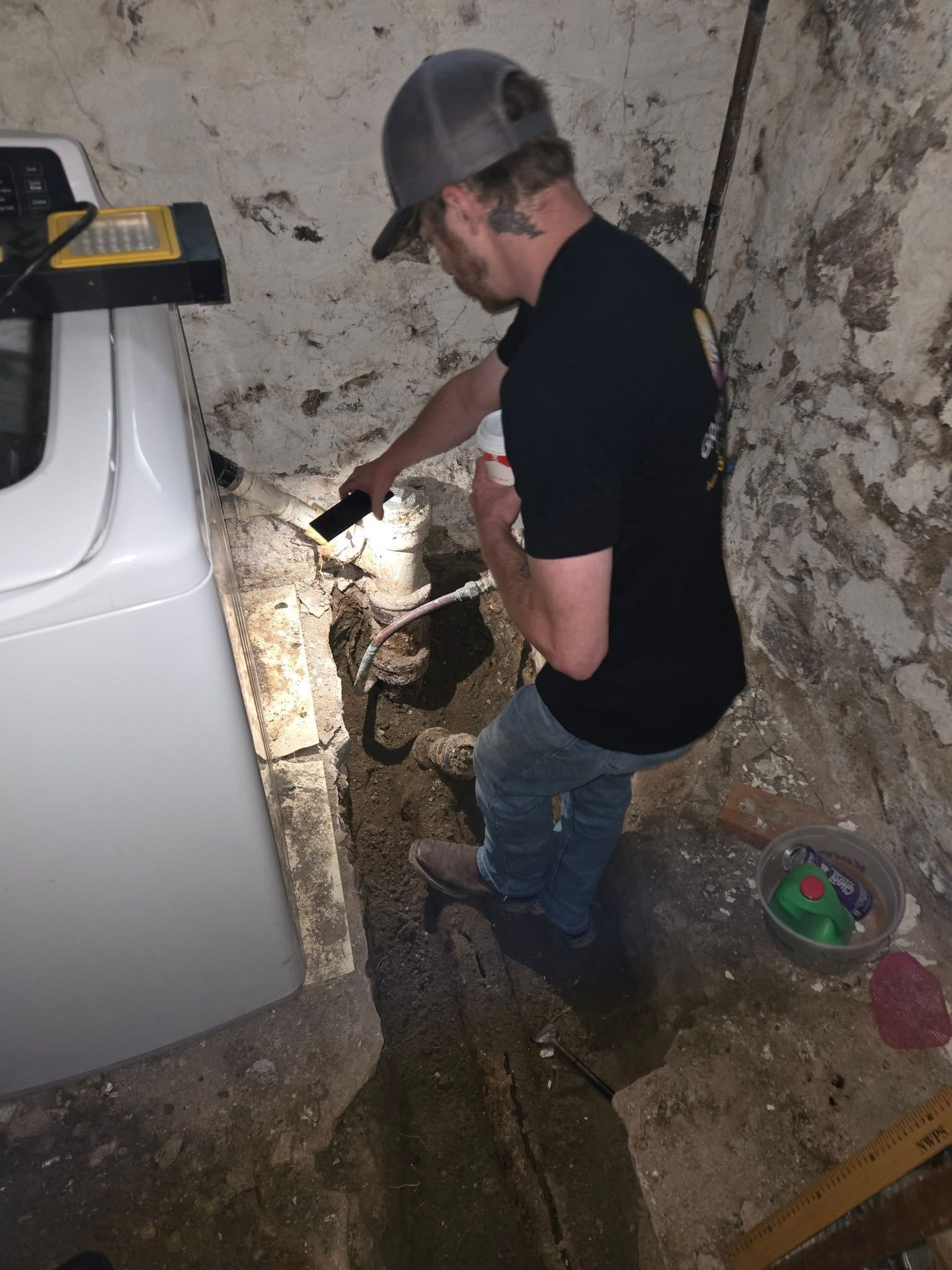 Man with flashlight inspects a basement wall near a washing machine. The basement is dirty with exposed stone.