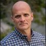 A bald man wearing a blue and white checkered shirt is looking at the camera.