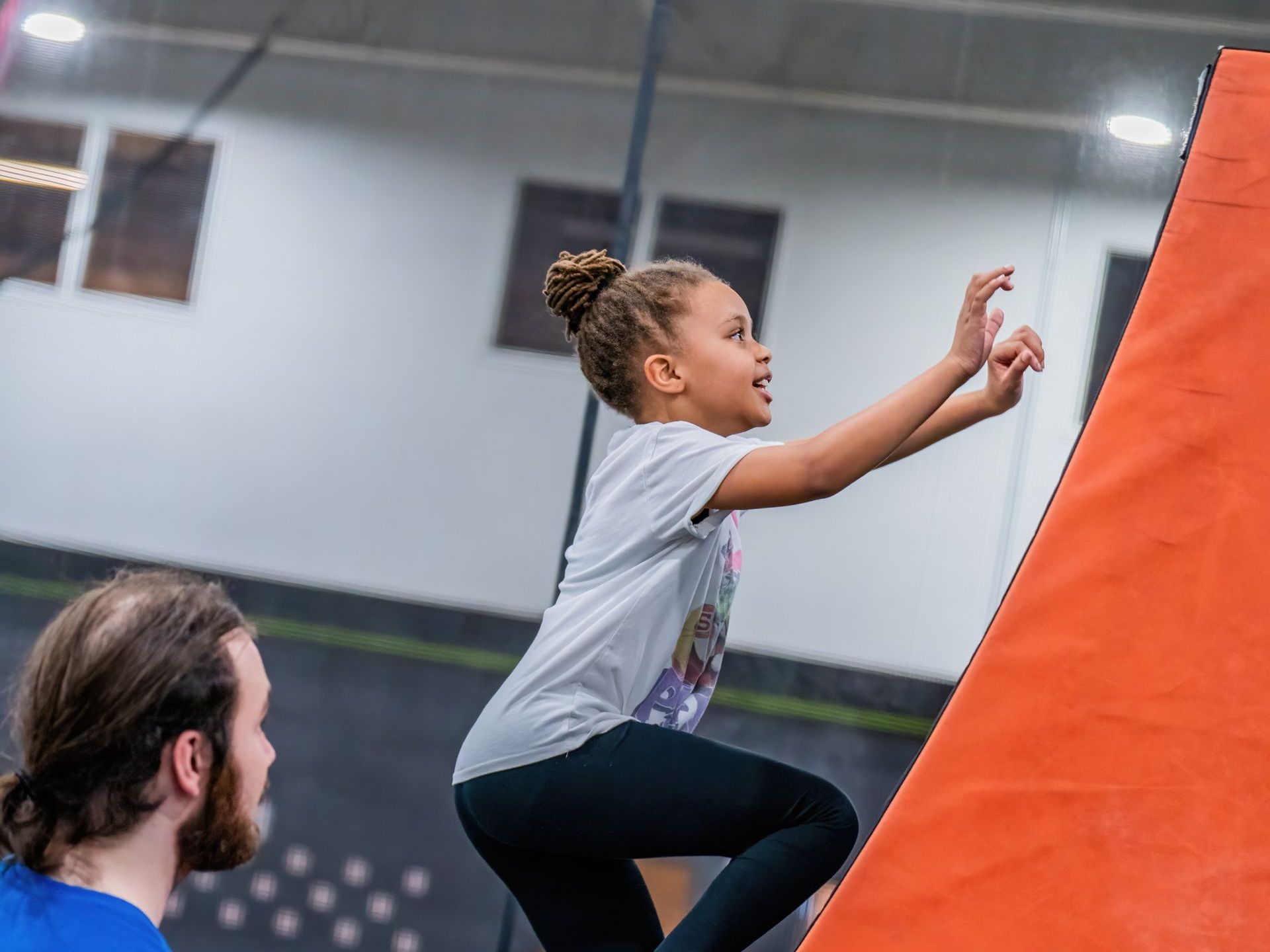 Girl climbing orange angled wall in an indoor recreation center, watched by a man in a blue shirt.