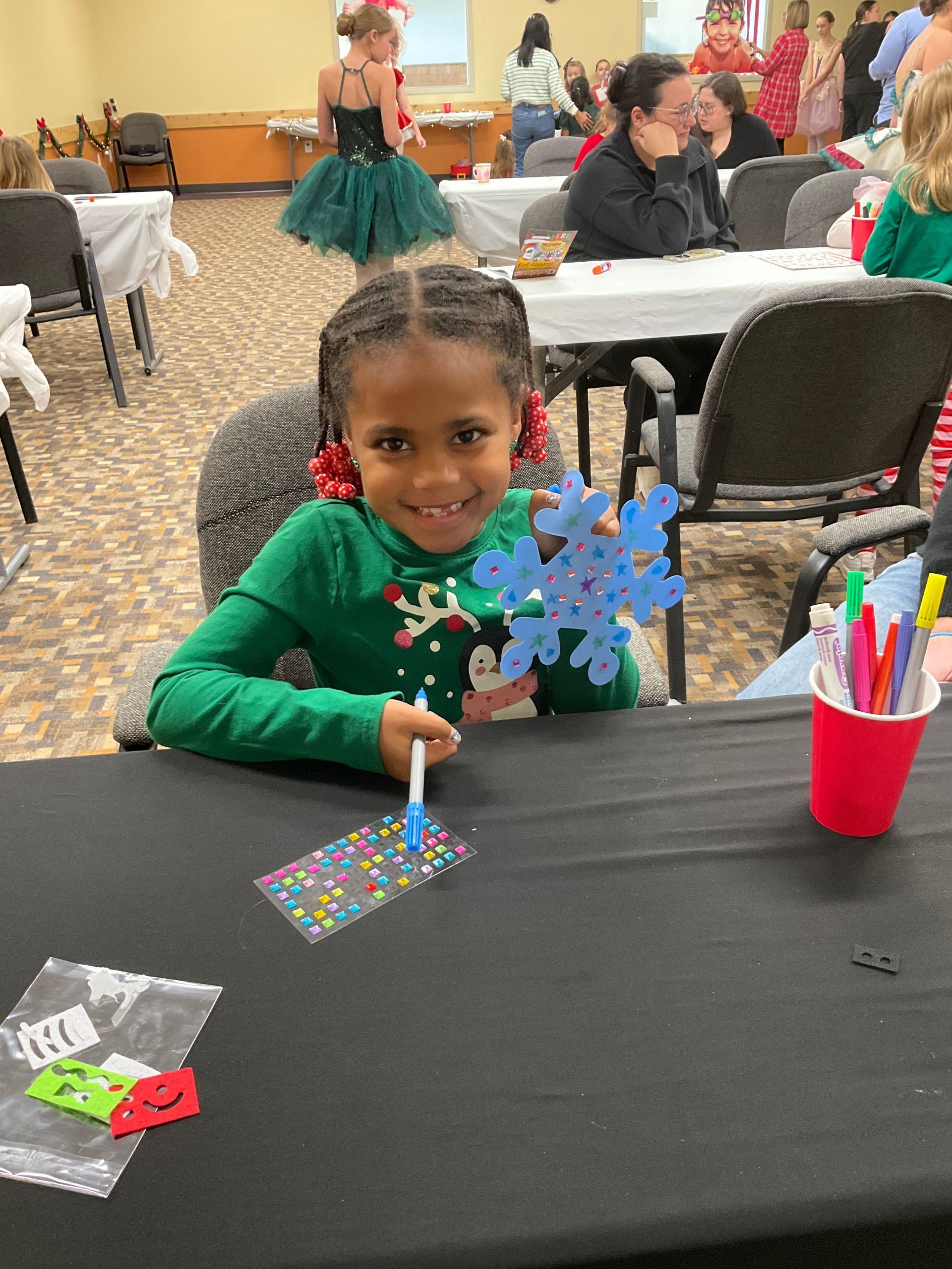 Young child at a table making crafts. She holds a blue snowflake decoration and smiles. A red cup with pens is visible.