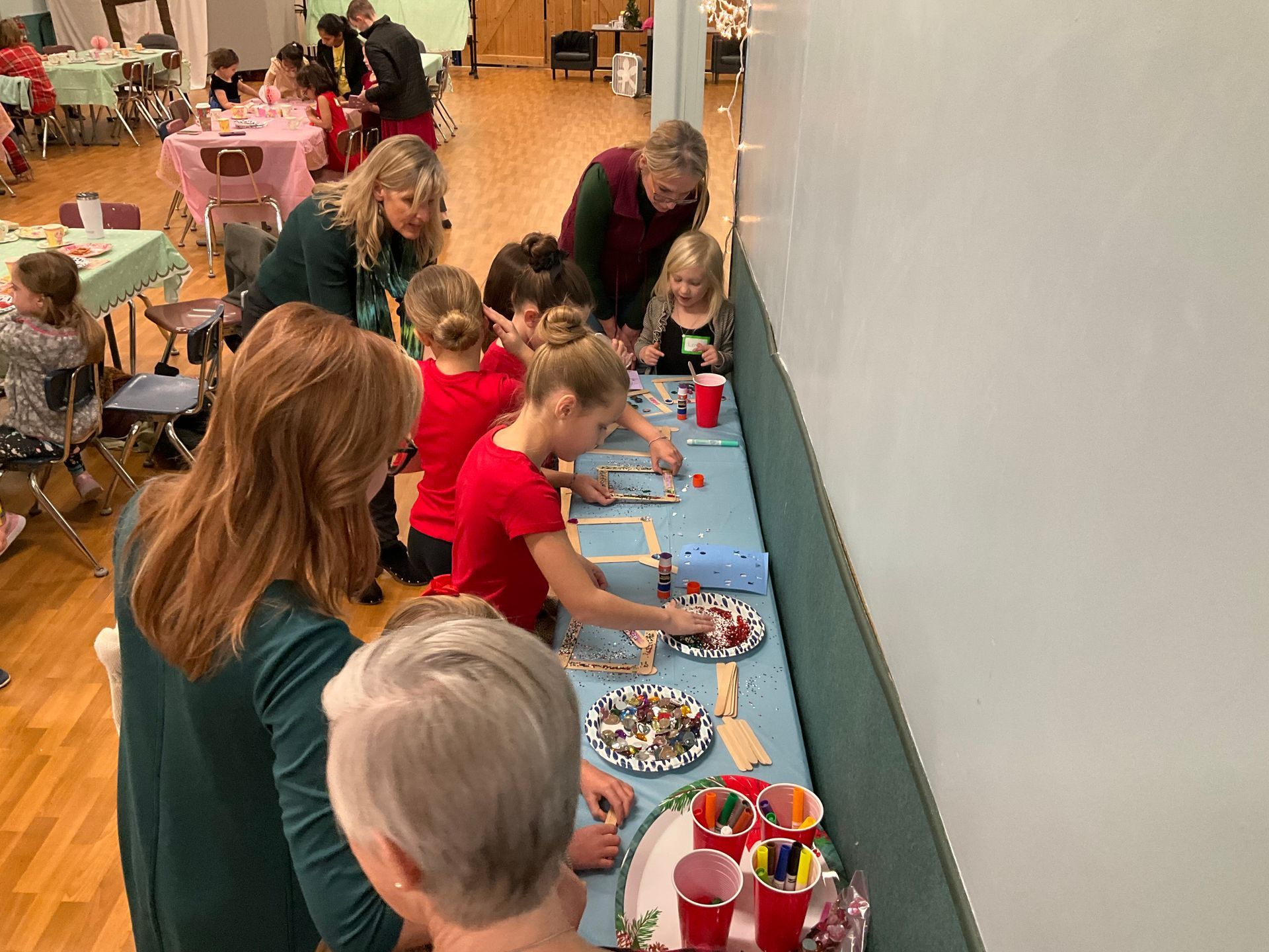 Children crafting at a table with adults assisting. Colorful crafts and supplies are visible in a brightly lit room.