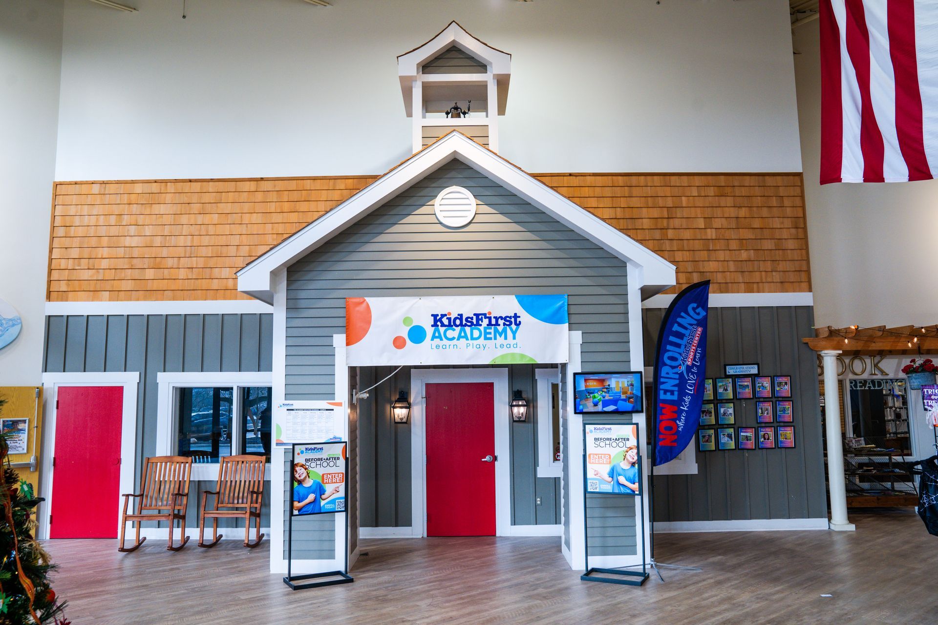 A miniature schoolhouse playset with a red door and banner, inside a building. A United States flag hangs on the right.