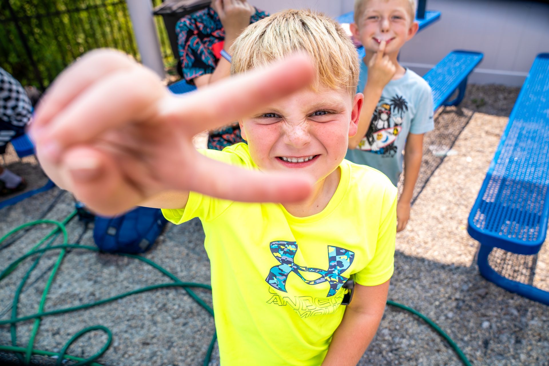 Boy in neon yellow shirt holds up peace sign toward camera, another boy behind him making a shushing gesture.